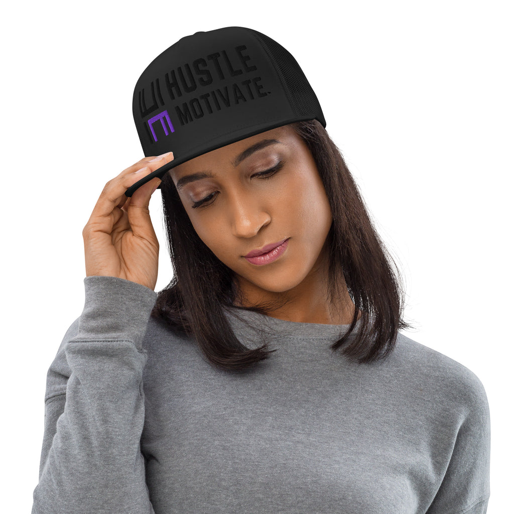 Model wearing the black Hustle Motivate Elevate trucker cap with tonal black lettering, a purple HM accent and black mesh panels