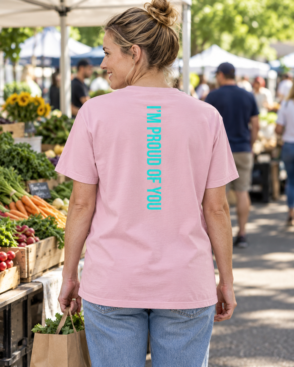 AI lifestyle mockup of the Blossom Hustle Motivate T-shirt worn at an outdoor market, showing the teal “I’M PROUD OF YOU” back design, alternate view.
